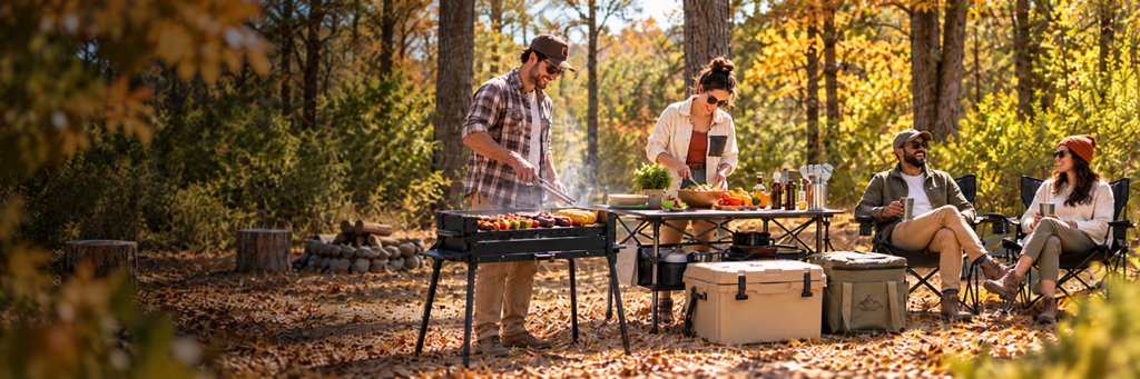 Group cooking on a portable charcoal grill with a rotomolded cooler and folding chairs at an autumn campsite.